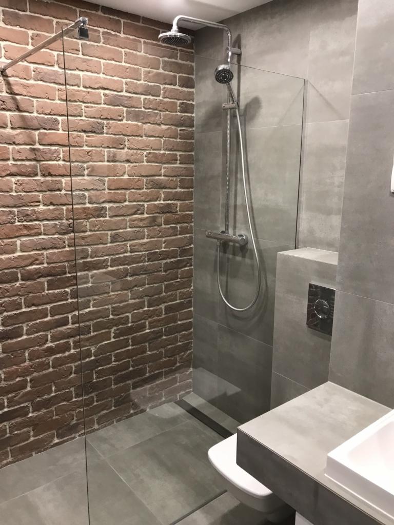 Nowoczesna łazienka z prysznicem typu walk-in, ściana wyłożona czerwoną cegłą, szara armatura i płytki imitujące beton.