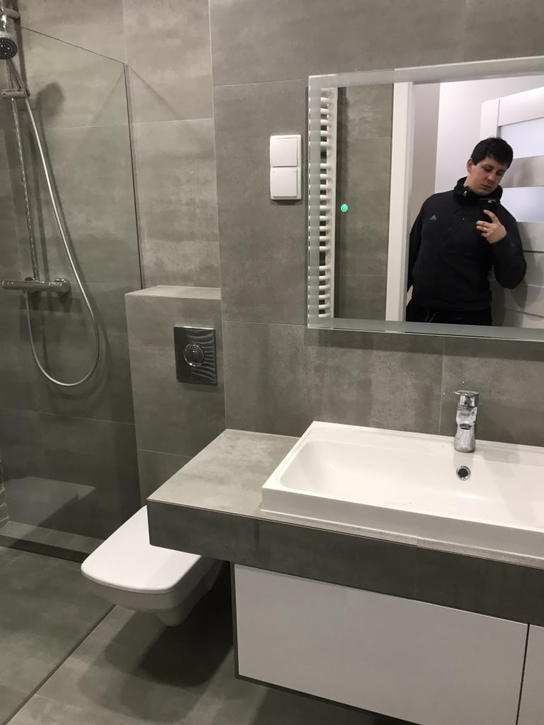 Nowoczesna łazienka w odcieniach szarości z białym umywalką i toaletą, widoczny prysznic za szklaną ścianką oraz odbicie osoby robiącej zdjęcie w podświetlanym lustrze.