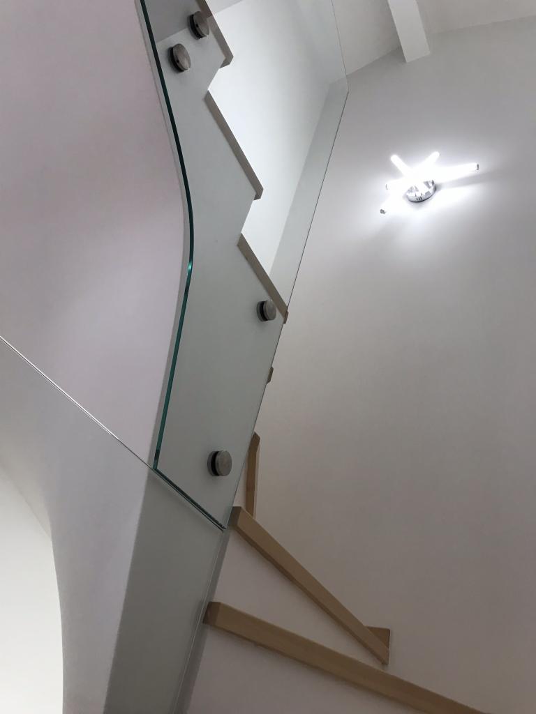 Nowoczesne schody z drewnianymi stopniami i szklaną balustradą mocowaną na metalowe uchwyty, oświetlone designerską lampą sufitową.