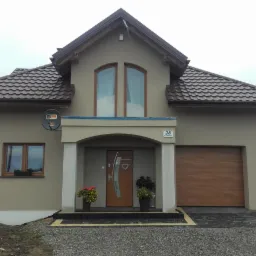 Nowoczesny dom jednorodzinny z brązowym dachem, beżową elewacją, drewnianą bramą garażową i drzwiami wejściowymi z elementami ze stali nierdzewnej, z numerem domu '28' nad wejściem.