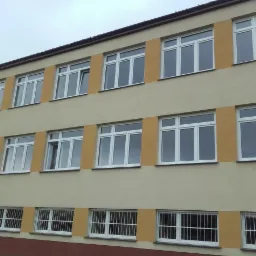 Elewacja budynku z trzema rzędami okien, dolny rząd z kratami, jedno okno otwarte na drugim piętrze, ściana w kolorze beżowym z pomarańczowymi wstawkami.