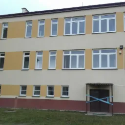 Fasada budynku z dwoma rzędami białych okien, dolne okna z kratami, wejście zabezpieczone taśmą.