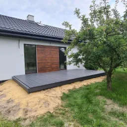 Nowoczesny dom z szarą elewacją i drewnianą deską elewacyjną przy tarasie z kompozytu w kolorze grafitu, na tle drzewa owocowego.