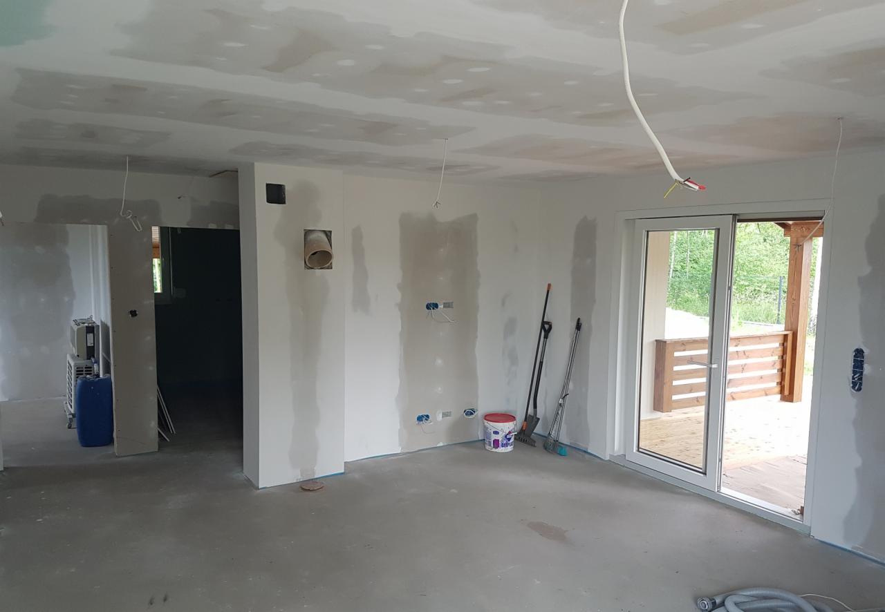 Wnętrze domu w trakcie wykańczania: ściany z widocznymi śladami szpachlowania, surowy betonowy podkład, przesuwne drzwi balkonowe z widokiem na drewniany taras, wystające kable elektryczne...