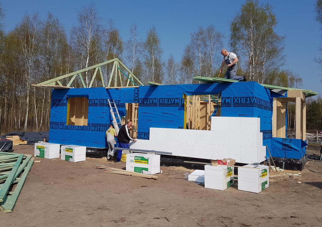 Budowa domu: pracownicy montują izolację styropianową na ścianach szkieletowej konstrukcji, zabezpieczonej niebieską membraną dachową Matrix Super, z widocznym szkieletem dachu i drewnianymi...