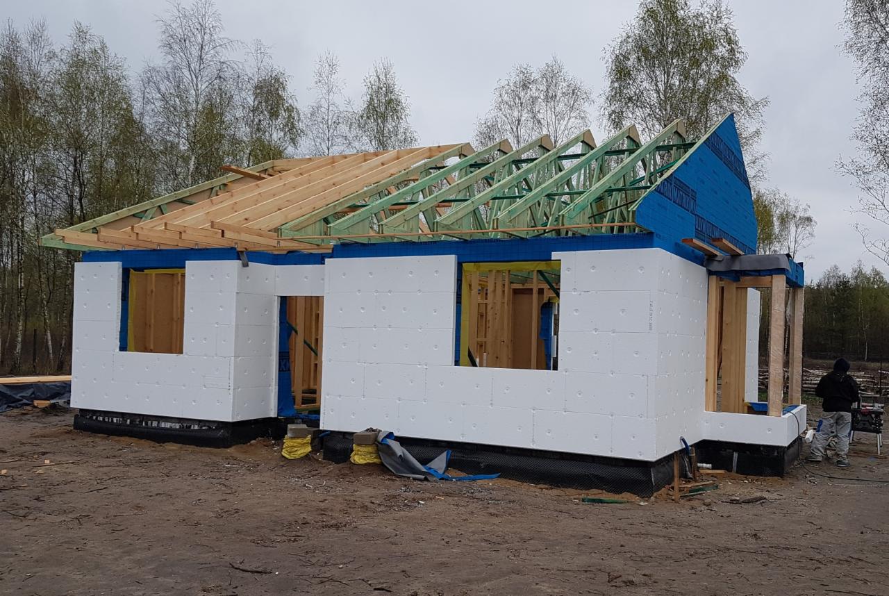 Budowa domu w technologii szkieletowej: Etap montażu izolacji termicznej ścian zewnętrznych z białego styropianu oraz konstrukcji dachu z drewnianych belek, widoczny pracownik w tle.