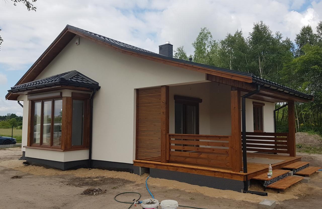 Parterowy dom szkieletowy z drewnianą werandą i oknem wykuszowym, wykończony w jasnej kolorystyce, z widocznymi puszkami farby na pierwszym planie.