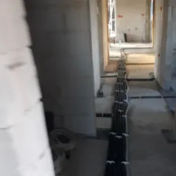 Wnętrze w stanie surowym z widocznymi przewodami elektrycznymi ułożonymi w korytku na podłodze, prowadzącymi przez kilka pomieszczeń.