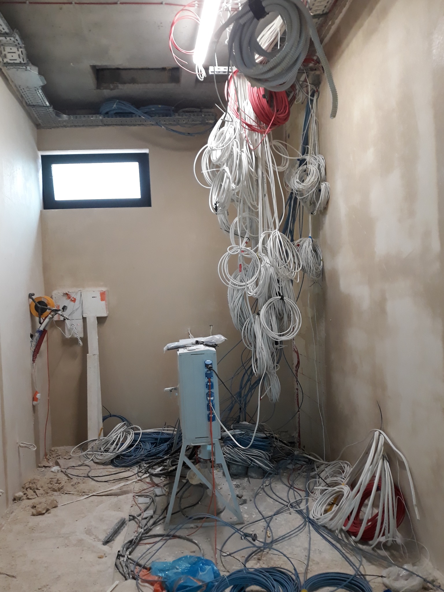 Wnętrze w trakcie prac elektrycznych: kable zwisające ze stropu, skrzynka elektryczna na statywie, narzędzia na ścianie, widoczne koryta kablowe pod sufitem, surowy stan wykończenia ścian.