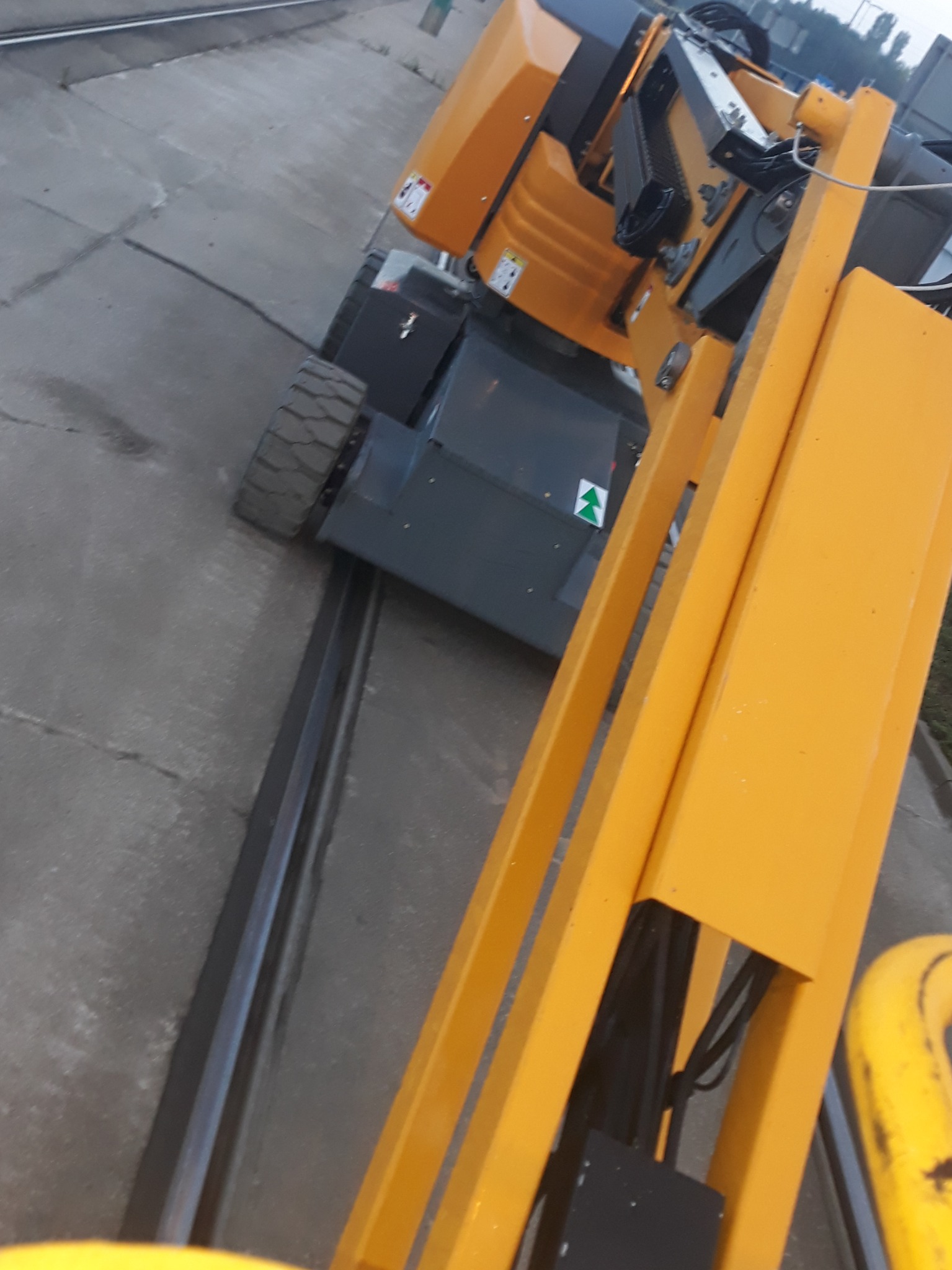 Żółty podnośnik koszowy na szynach, widok z góry, szare betonowe podłoże, pochmurne niebo w tle.