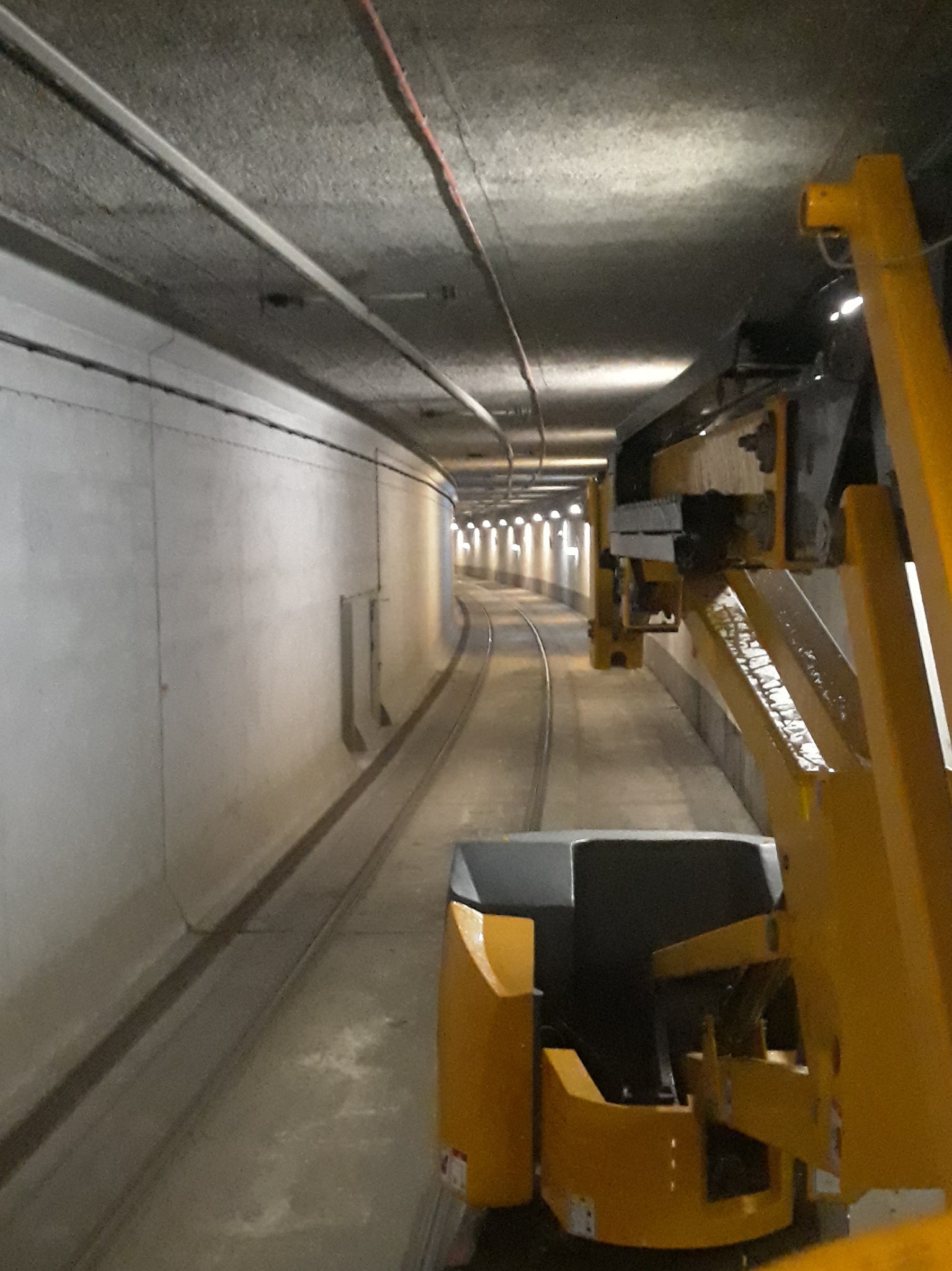 Wnętrze tunelu z szarą betonową konstrukcją i oświetleniem w oddali, widok z żółtego pojazdu szynowego z elementami hydraulicznymi.
