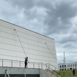 Wash Complex - Pracownicy z długimi lancami czyszczą elewację budynku z poziomu podestu i schodów. Widoczne okna i biała, panelowa fasada budynku w pochmurny dzień.