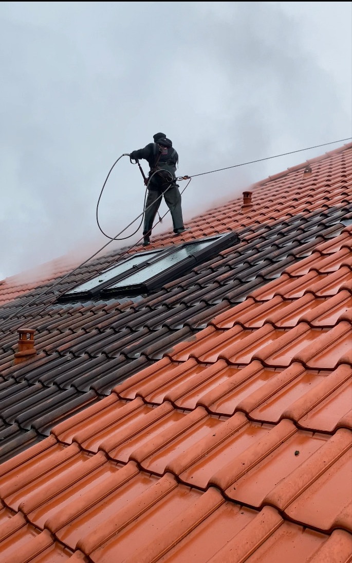 Czyszczenie dachu z dachówki ceramicznej myjką ciśnieniową. Widoczna różnica w kolorze między umytą a brudną częścią dachu. Praca na wysokości.