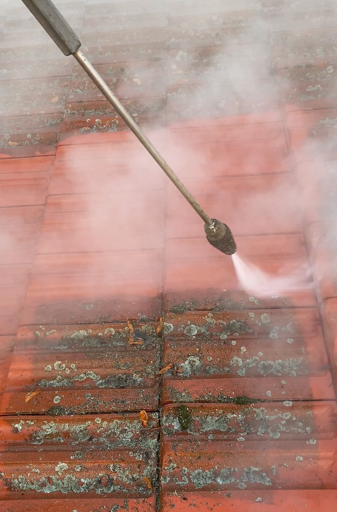 Czerwona dachówka czyszczona lancą ciśnieniową, z widocznymi smugami po usunięciu zabrudzeń i mchu, otoczona parą wodną.