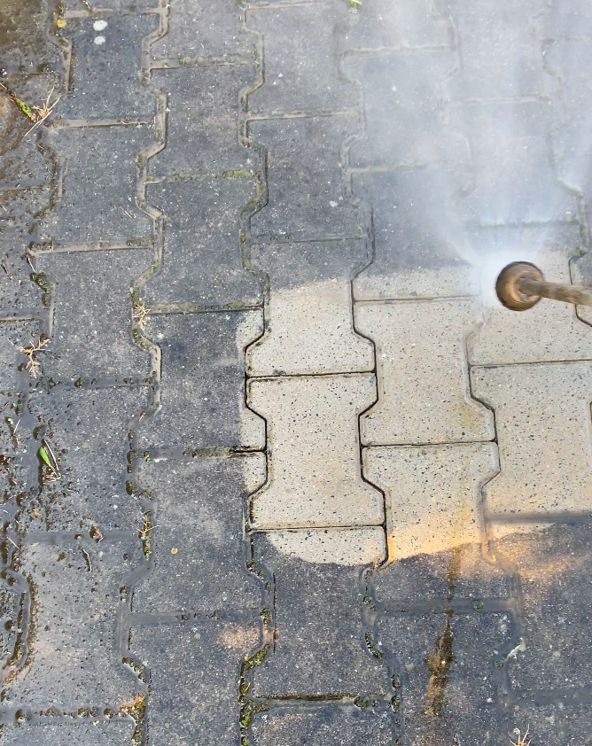 Mycie kostki brukowej: połowa wyczyszczona, widoczna różnica w kolorze i czystości. Dysza ciśnieniowa w akcji, woda spływa po powierzchni.