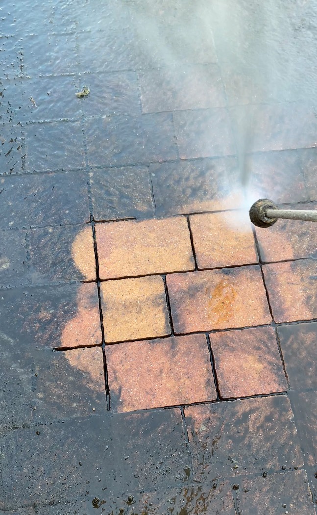 Ceglana kostka brukowa czyszczona myjką ciśnieniową, widoczna różnica w kolorze między czystą a zabrudzoną powierzchnią, strumień wody pod dużym ciśnieniem.
