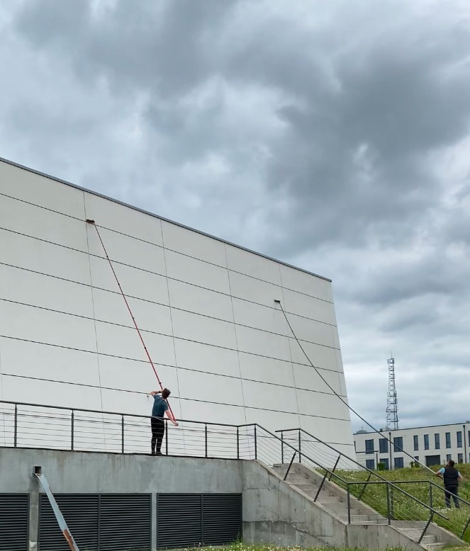 Pracownicy z długimi lancami czyszczą elewację budynku z poziomu podestu i schodów. Widoczne okna i biała, panelowa fasada budynku w pochmurny dzień.