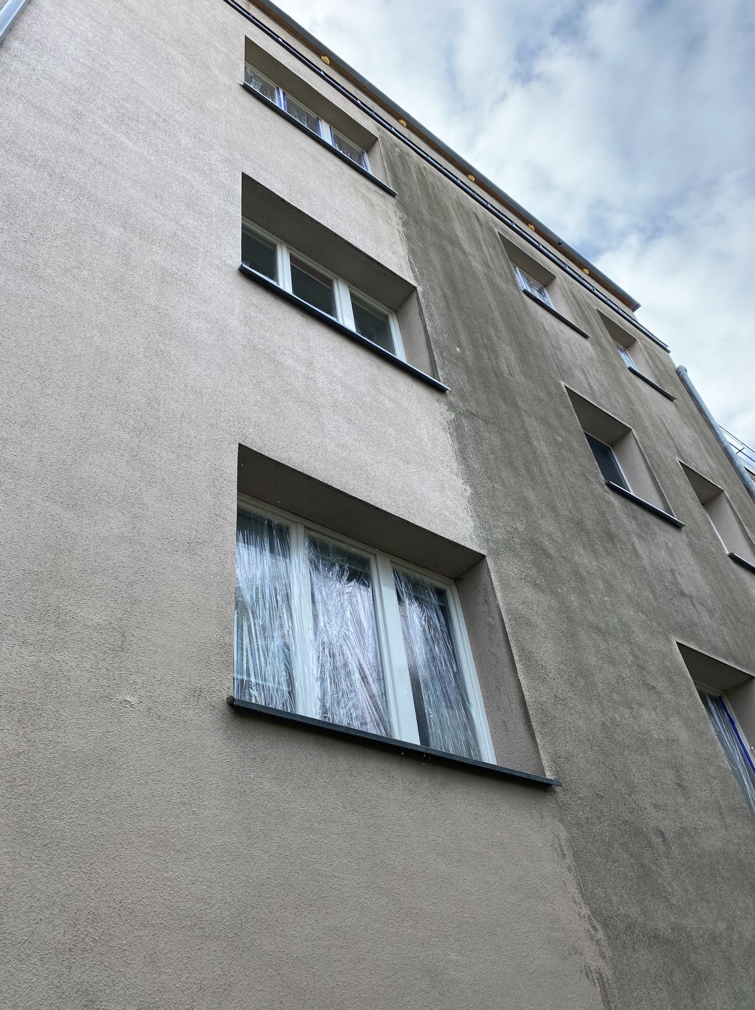 Elewacja budynku przed czyszczeniem: widoczne zabrudzenia i okna zabezpieczone folią. Perspektywa z dołu, pochmurne niebo w tle.