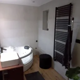 Nowoczesna łazienka z narożną wanną z hydromasażem, umywalką nablatową na brązowej szafce, czarnym grzejnikiem drabinkowym z wiszącym ręcznikiem i drewnopodobną podłogą.