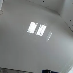 Wylany, samopoziomujący podkład podłogowy w trakcie schnięcia, z widocznymi refleksami światła z okna, obok wiadro z narzędziami.