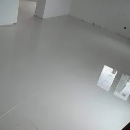 Błyszcząca, jasna podłoga z płytek ceramicznych w trakcie układania, widoczne ślady po zaprawie i niedoczyszczone fugi, fragment ściany z niedokończonymi pracami wykończeniowymi.