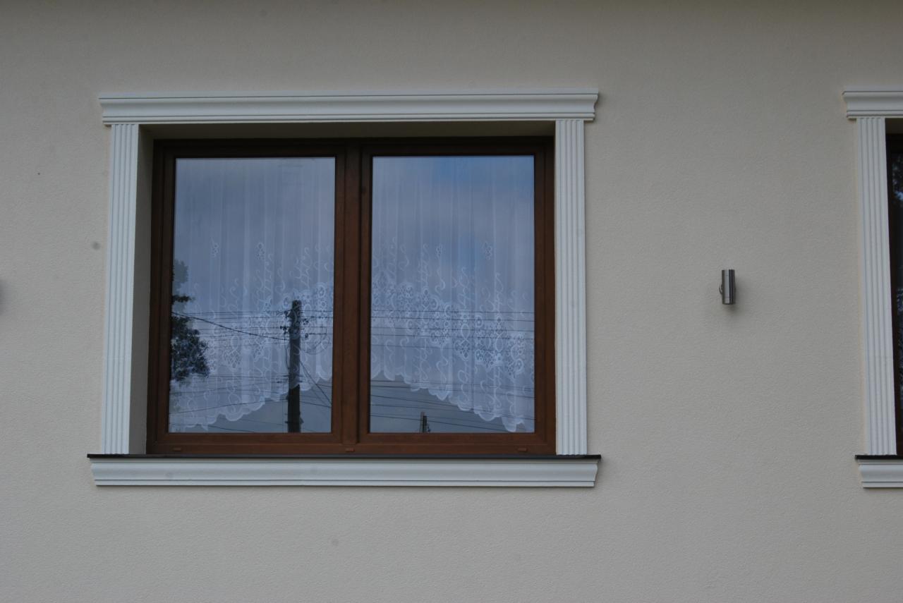 Elewacja budynku z dwoma oknami z brązowymi ramami i białymi, dekoracyjnymi obramowaniami, widoczna faktura tynku i mała lampa ścienna.