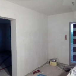 Świeżo otynkowane ściany w pomieszczeniu w trakcie remontu, widoczny otwór drzwiowy, kable na podłodze i narzędzia pracy przy drzwiach.