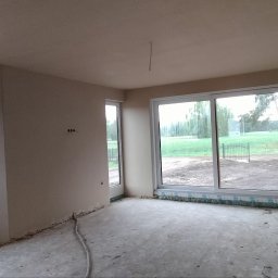 PIOTR - BUD PIOTR BIESALSKI - Wnętrze po tynkowaniu maszynowym. Widoczne gładkie ściany, okna z widokiem na zieleń i surowa podłoga. Przygotowane punkty elektryczne na ścianach.