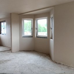 PIOTR - BUD PIOTR BIESALSKI - Wnętrze po tynkowaniu maszynowym: jasne ściany, okna z widokiem na zieleń, widoczne instalacje elektryczne i rury. Stan surowy przed wykończeniem.