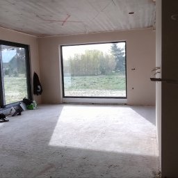 PIOTR - BUD PIOTR BIESALSKI - Wnętrze w stanie surowym: ściany otynkowane, duże okna z widokiem na zieleń, widoczne instalacje elektryczne i betonowa podłoga. Ślady prac remontowych.