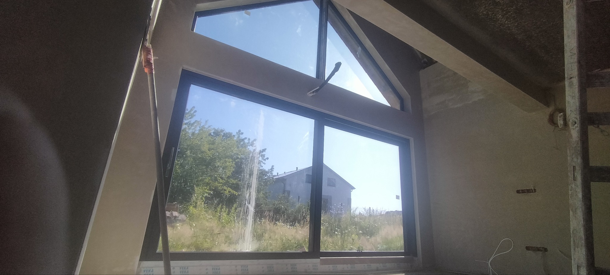Wnętrze po tynkowaniu maszynowym z dużym oknem, widok na zewnątrz. Ściany jasne, surowe wykończenie, widoczne elementy konstrukcyjne dachu.