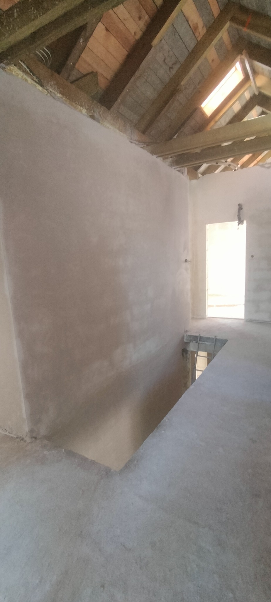 Wnętrze domu w trakcie remontu: świeżo otynkowane ściany poddasza z drewnianą konstrukcją dachu i oknem dachowym, surowy beton na podłodze i otwór w stropie.