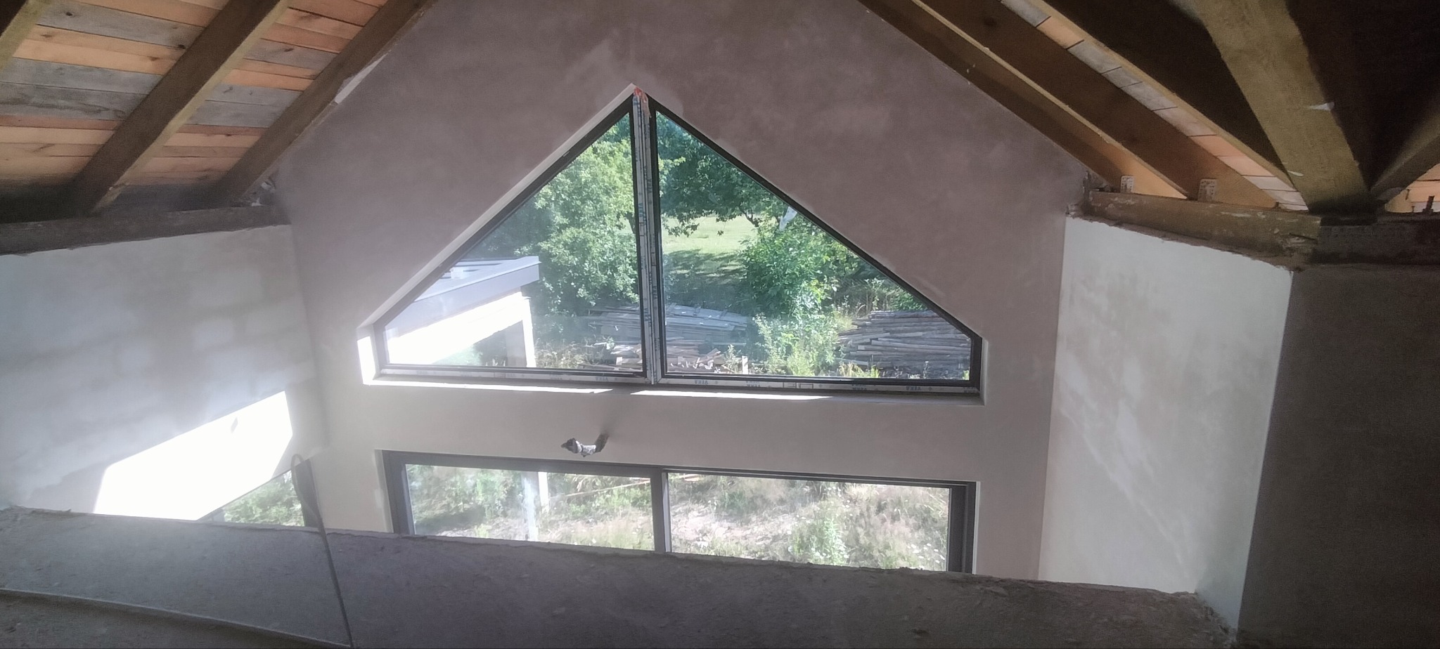 Wnętrze z oknami o nietypowych kształtach po tynkowaniu ścian. Widoczne drewniane elementy konstrukcyjne dachu. Świeżo położony tynk o jasnoszarym odcieniu.