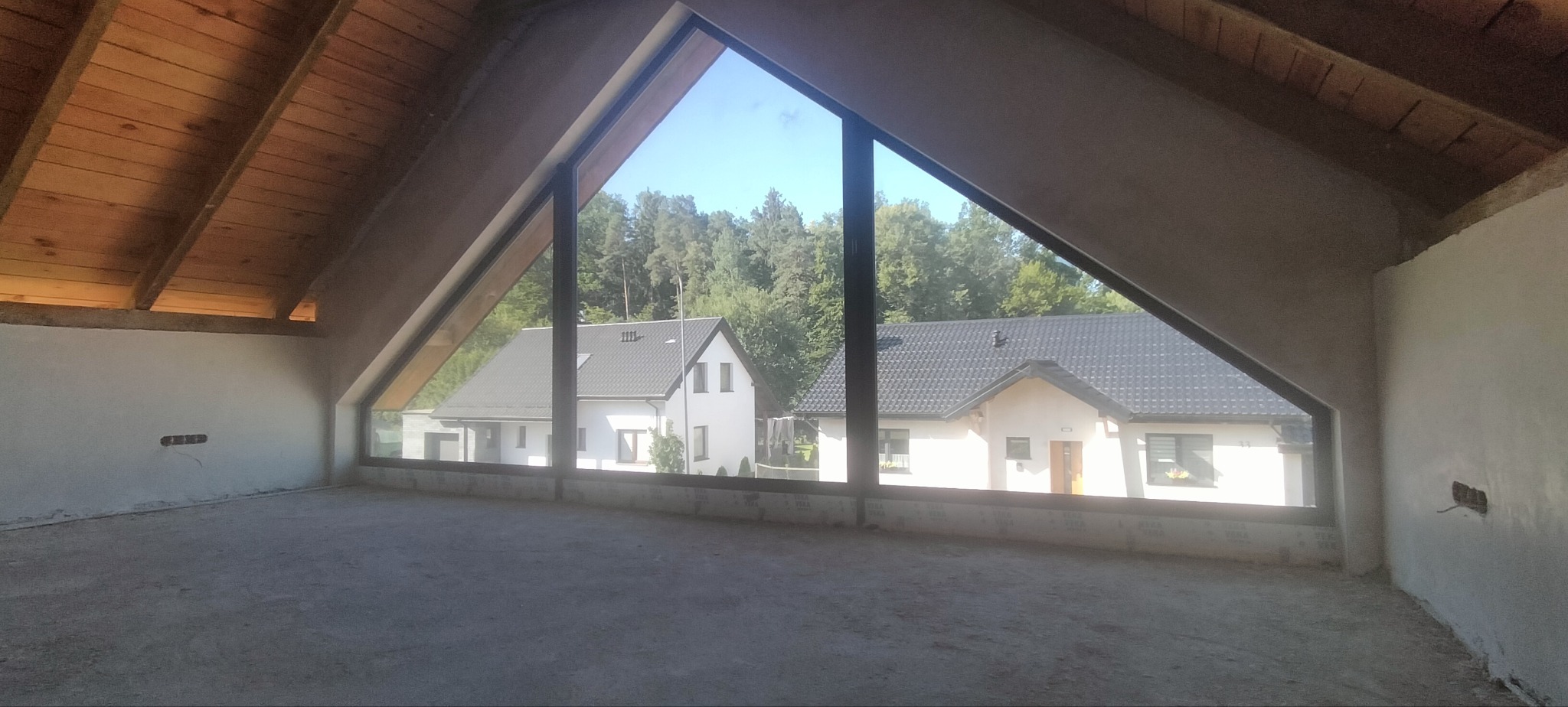 Poddasze w trakcie prac wykończeniowych: surowe ściany, drewniana więźba dachowa i widok na domy przez trójkątne okno. Wnętrze do adaptacji.