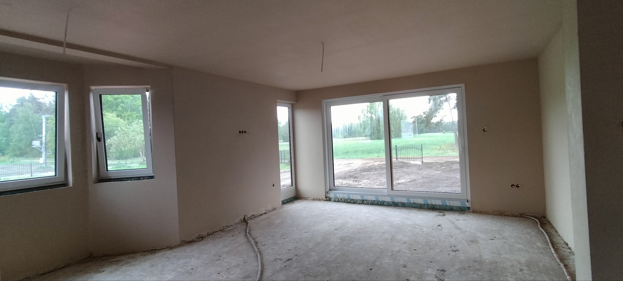 Wnętrze po tynkowaniu maszynowym. Widoczne gładkie ściany, okna z widokiem na zieleń i surowa podłoga. Przygotowane punkty elektryczne na ścianach.