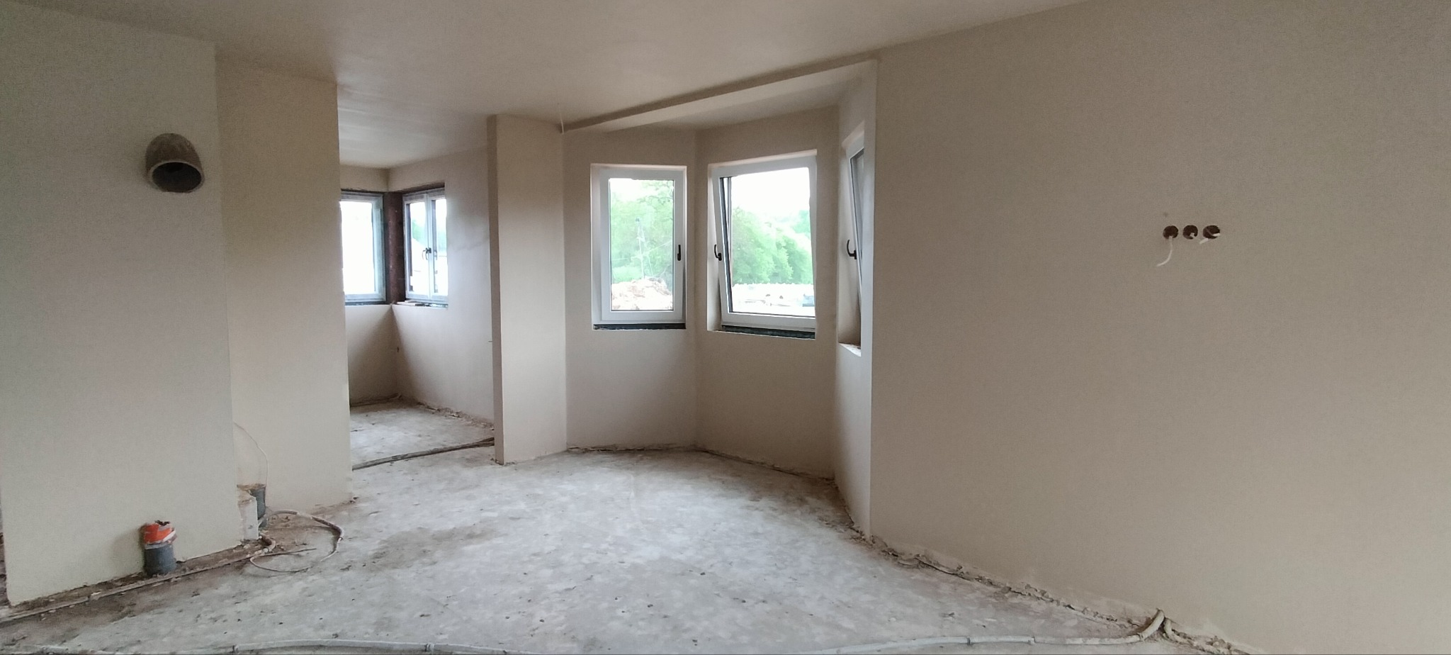 Wnętrze po tynkowaniu maszynowym: jasne ściany, okna z widokiem na zieleń, widoczne instalacje elektryczne i rury. Stan surowy przed wykończeniem.