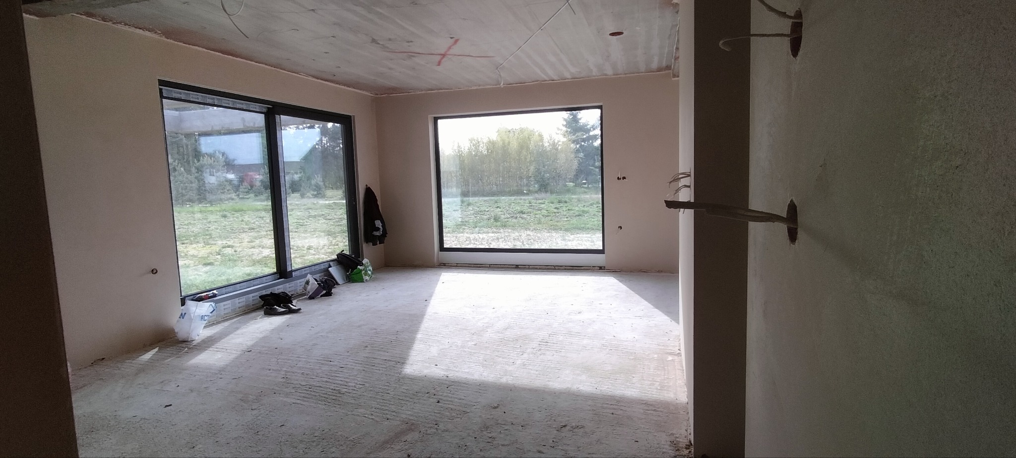 Wnętrze w stanie surowym: ściany otynkowane, duże okna z widokiem na zieleń, widoczne instalacje elektryczne i betonowa podłoga. Ślady prac remontowych.