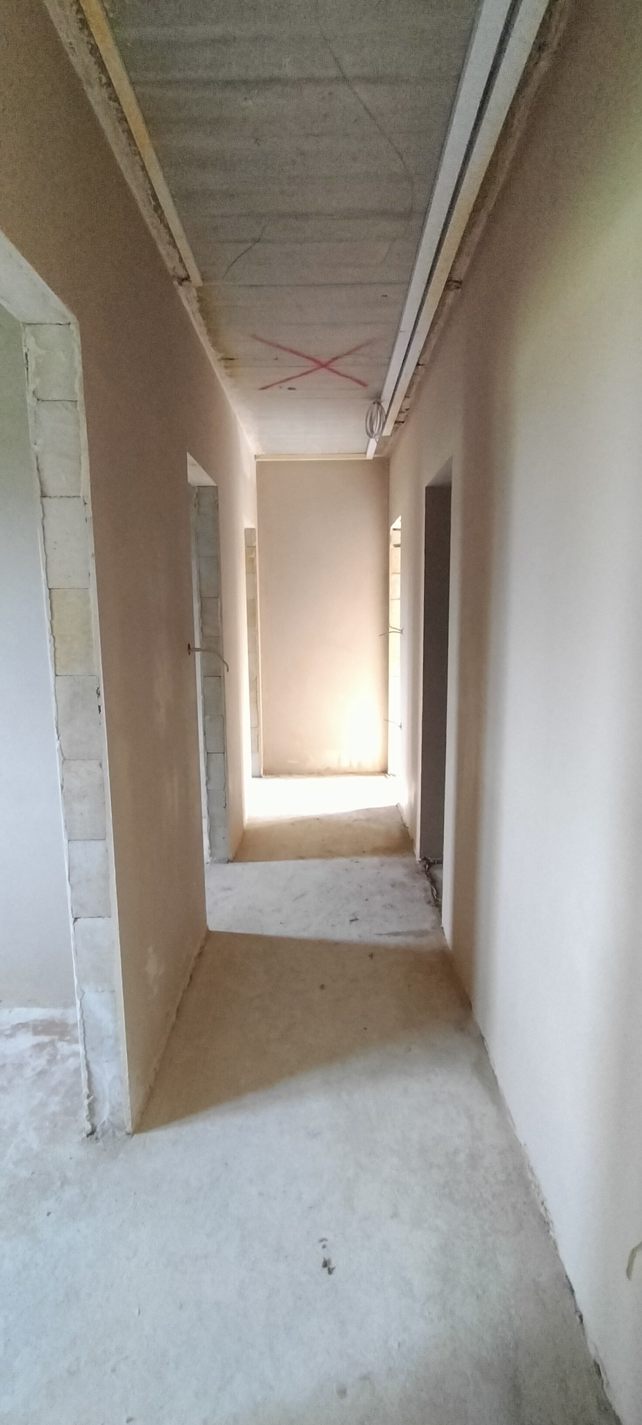 Wnętrze w trakcie prac wykończeniowych: świeżo otynkowane ściany w jasnym odcieniu beżu, surowy betonowy sufit z widocznymi oznaczeniami krzyżowymi i otworami drzwiowymi.