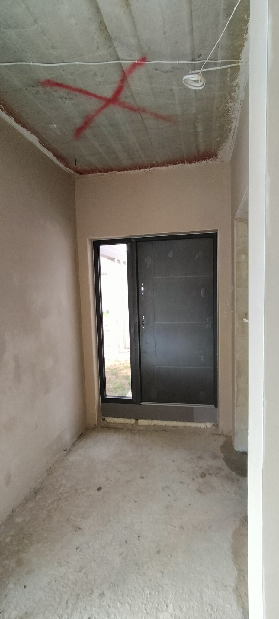 Wnętrze w trakcie wykończenia: ściany po tynkowaniu, surowy sufit z instalacją i czerwoną farbą, widoczne drzwi wejściowe. Etap prac wykończeniowych.