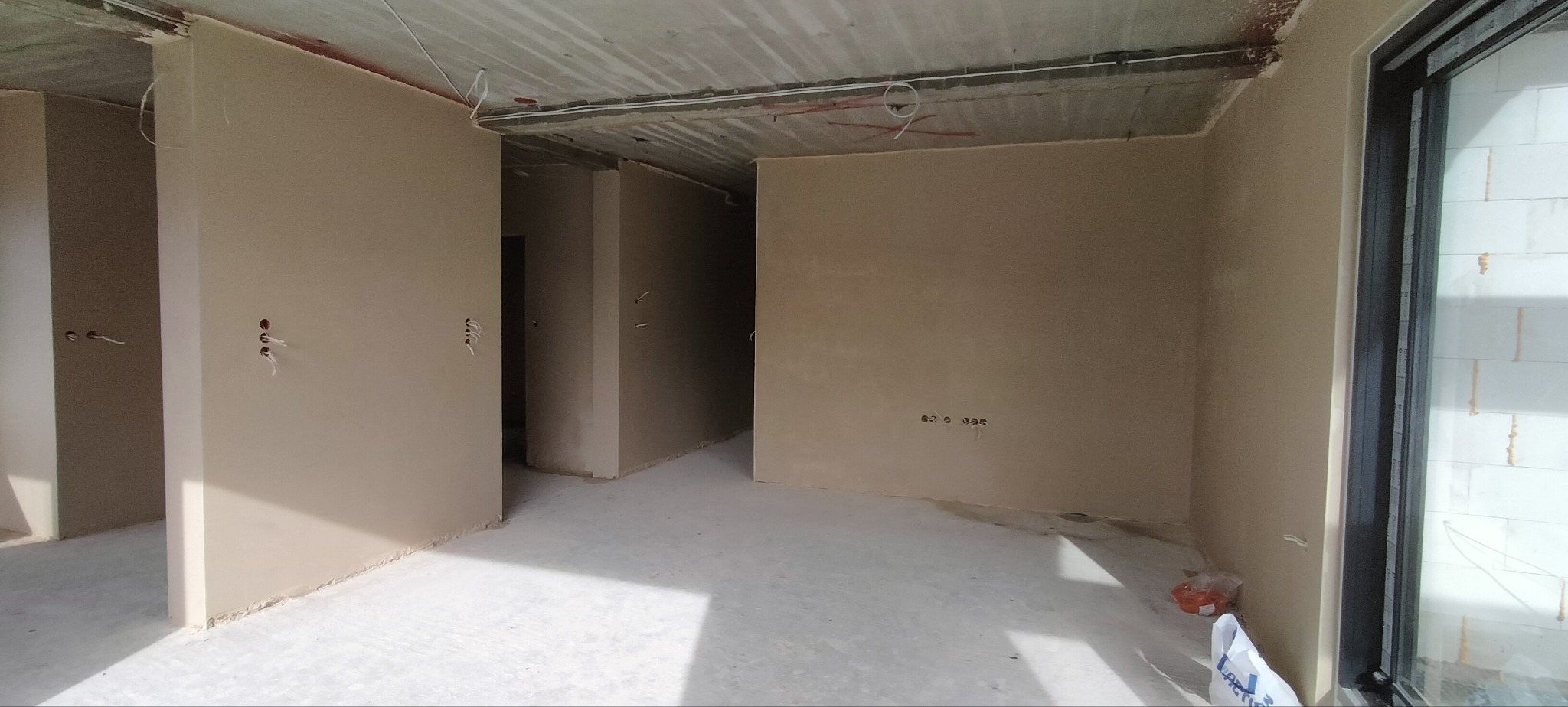 Wnętrze po tynkowaniu maszynowym: gładkie, beżowe ściany, surowy betonowy sufit z instalacjami. Widoczne otwory pod gniazdka i przewody.