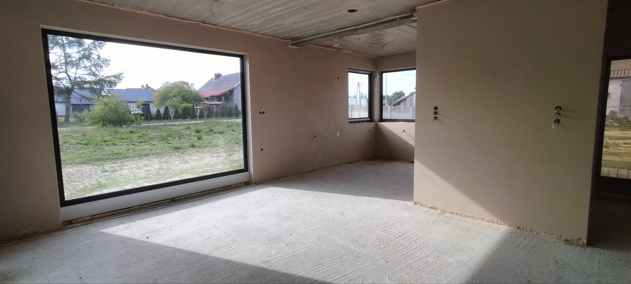 Wnętrze domu po tynkowaniu maszynowym. Widoczne duże okno z widokiem na zewnątrz, ściany z gładzią i instalacje elektryczne. Surowy stan wykończenia.