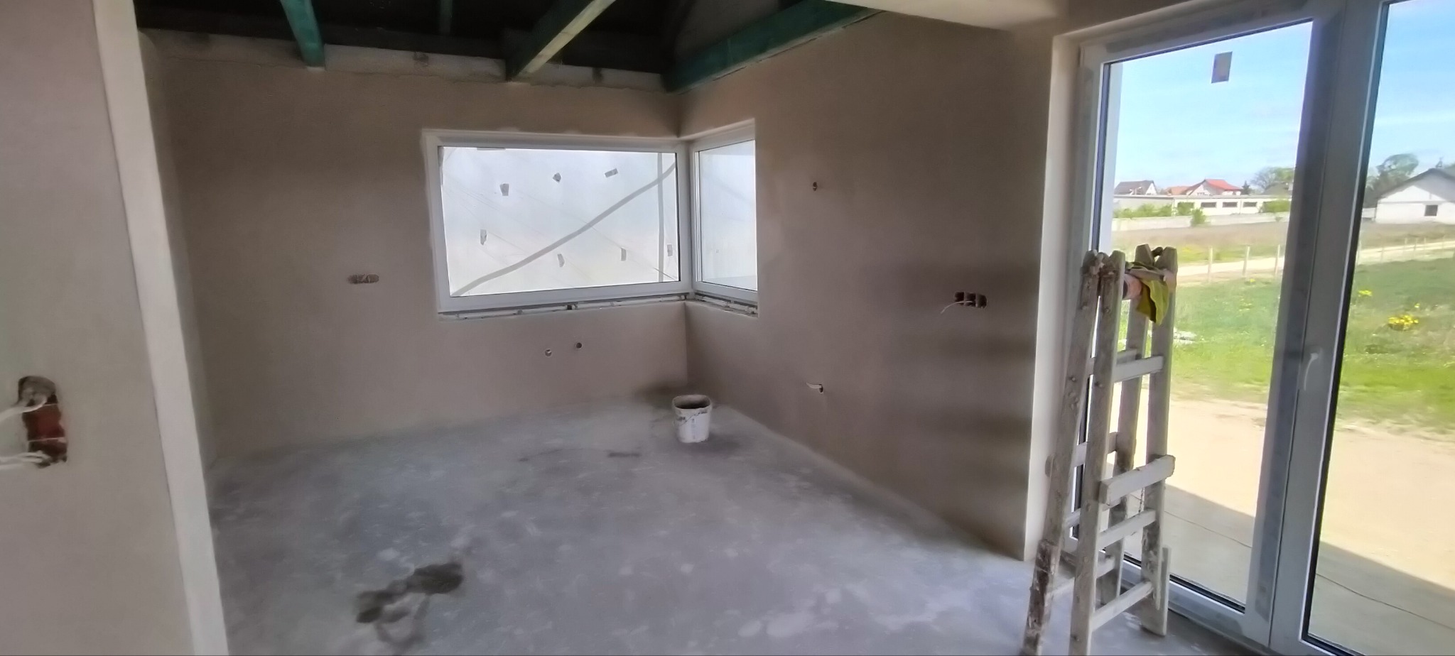 Wnętrze po tynkowaniu maszynowym: gładkie ściany, okna zabezpieczone folią, drabina oparta o ścianę. Widoczne puszki elektryczne i wiadro. Jasne pomieszczenie.