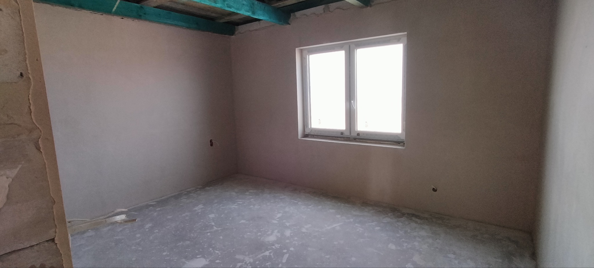 Pokój po tynkowaniu maszynowym. Ściany gładkie, okno białe, sufit z drewnianymi belkami pomalowanymi na zielono. Podłoga przygotowana pod wylewkę. Widoczne gniazdka.