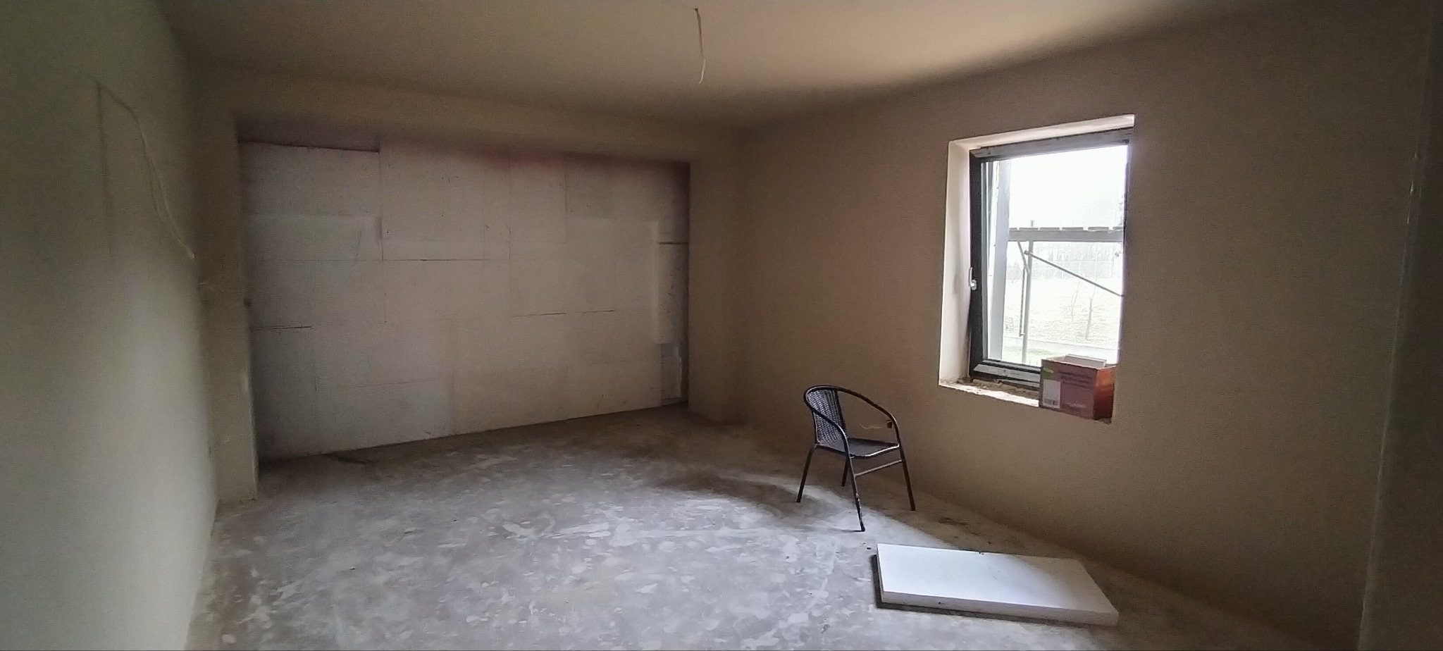 Surowe wnętrze pokoju z oknem, krzesłem i częściowo otynkowanymi ścianami. Widoczny etap prac wykończeniowych w nowym budynku lub podczas remontu.