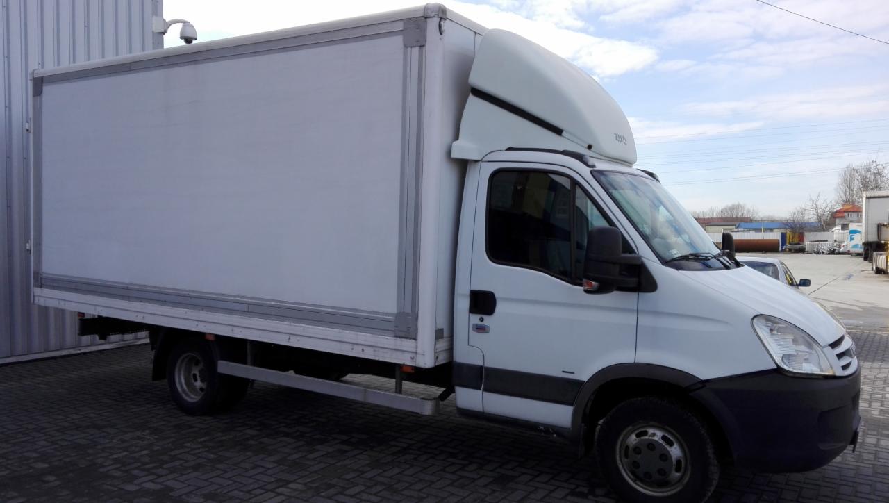 Biały samochód dostawczy typu furgon z zabudową kontenerową na tle parkingu z kostki brukowej i budynków przemysłowych w oddali.