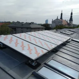 Rząd paneli słonecznych marki Viessmann na metalowym dachu, z widokiem na budynki miejskie i kościół z wieżami w tle, pochmurne niebo.