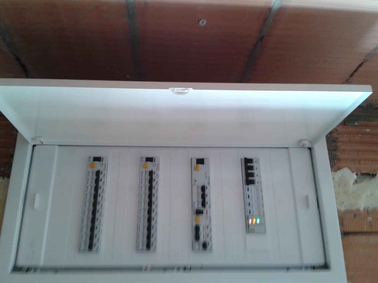 Otwarta skrzynka rozdzielcza z czterema modułami zabezpieczeń, widoczne przewody i diody sygnalizacyjne, surowy mur w tle.