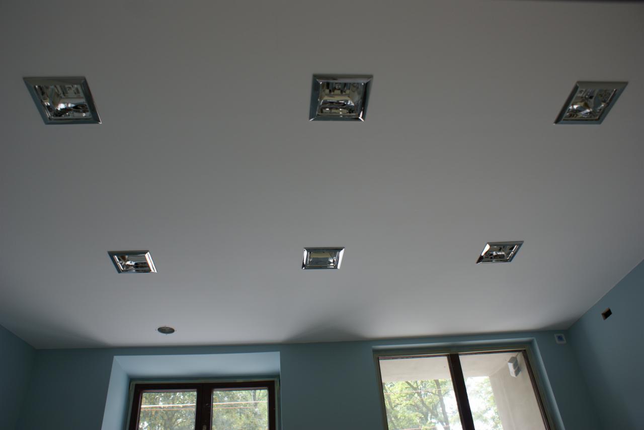 Sześć kwadratowych lamp sufitowych z metalowymi obramowaniami w białym suficie, widok z dołu, okna z widokiem na drzewa w tle.