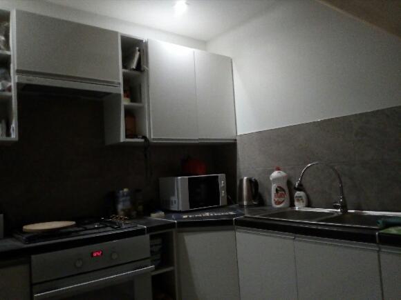 Wyposażona kuchnia z białymi szafkami wiszącymi i stojącymi, szarym blatem, kuchenką i zlewem, widok na fragment ściany z szarą mozaiką.