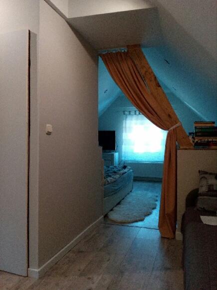 Adaptacja poddasza w Obornikach Śląskich: widok na sypialnię z łóżkiem, dywanem i zasłoną oddzielającą przestrzeń, widoczne skośne ściany.